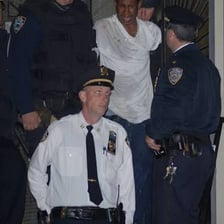 A handcuffed suspect being escorted by police past waiting cameras