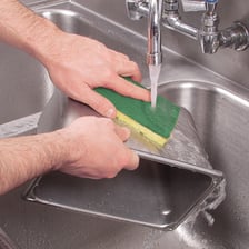 Hands scrubbing a kitchen sink with a soapy scouring pad