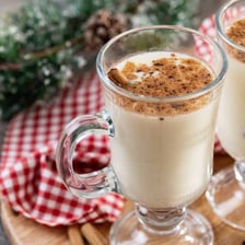 A glass of eggnog garnished with nutmeg beside Christmas decorations.