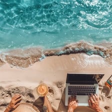 A writer using a laptop on a sunny beach beside sunscreen