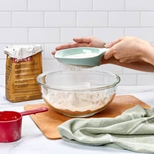 A handheld sifter being used to sift flour into a bowl.