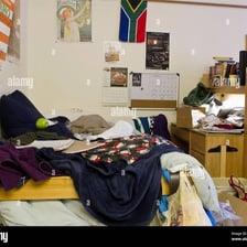 A cluttered college dorm room with clothes and belongings scattered around.