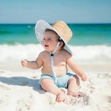 A baby wearing a brimmed sun hat outdoors at a beach.