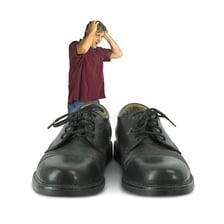 A person beside oversized shoes illustrating the phrase “big shoes to fill.”