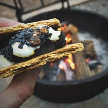 A toasted marshmallow and chocolate sandwiched between graham crackers beside a campfire