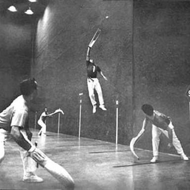 A jai alai player uses a curved cesta to throw a ball against a fronton wall.