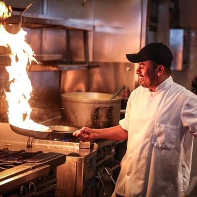 A cook flambés food in a pan, igniting alcohol to create a brief flame.