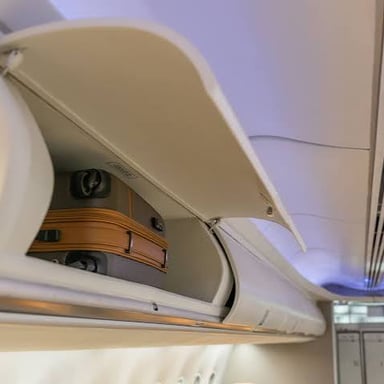 An open overhead compartment above airplane seats, with carry-on luggage inside.
