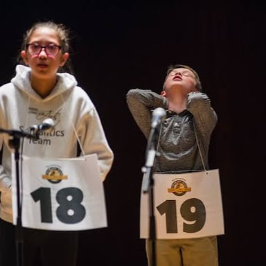A contestant standing at a spelling bee and spelling a word aloud.