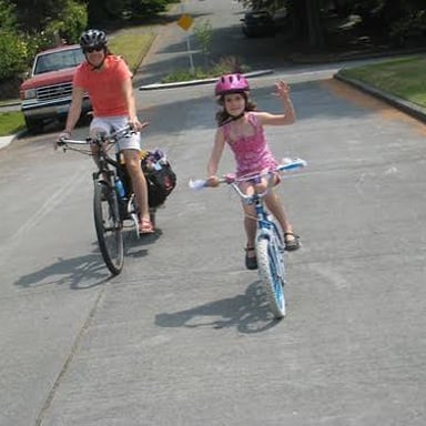 A child rides a bicycle without holding the handlebars while a parent watches.