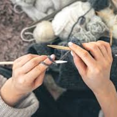 Hands using knitting needles to form fabric from yarn.